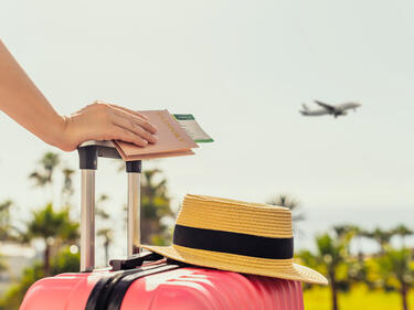 Woman with pink suitcase and passport with boarding pass standing on passengers ladder of airplane opposite sea with palm trees.