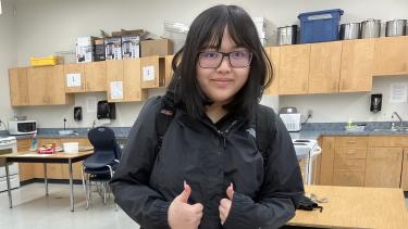 Grade 12 female student with their thumbs up, standing in front of food supplies